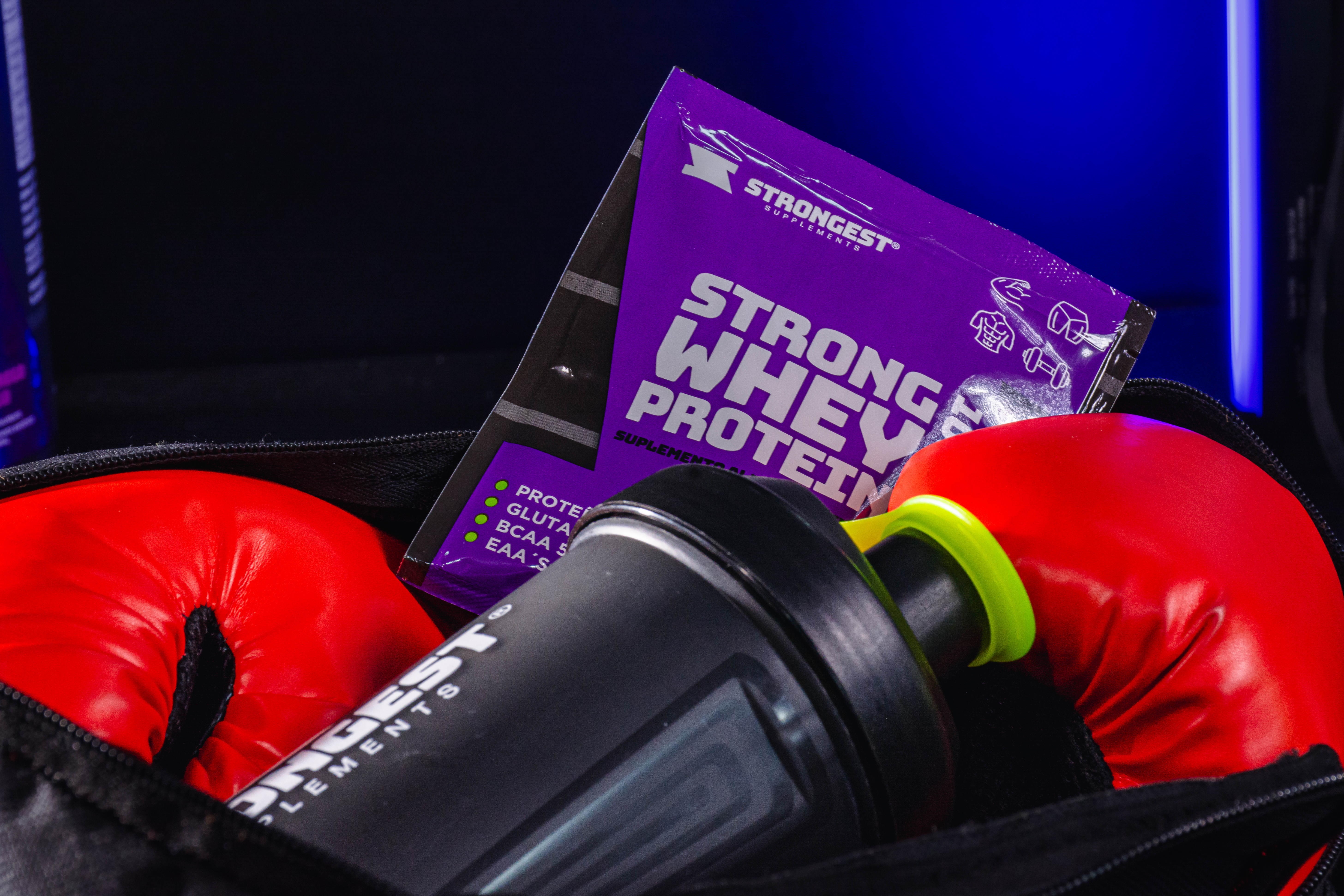 Sachê do Strong Whey Protein da Strongest dentro de uma bolsa de academia, ao lado de luvas de boxe vermelhas e um shaker