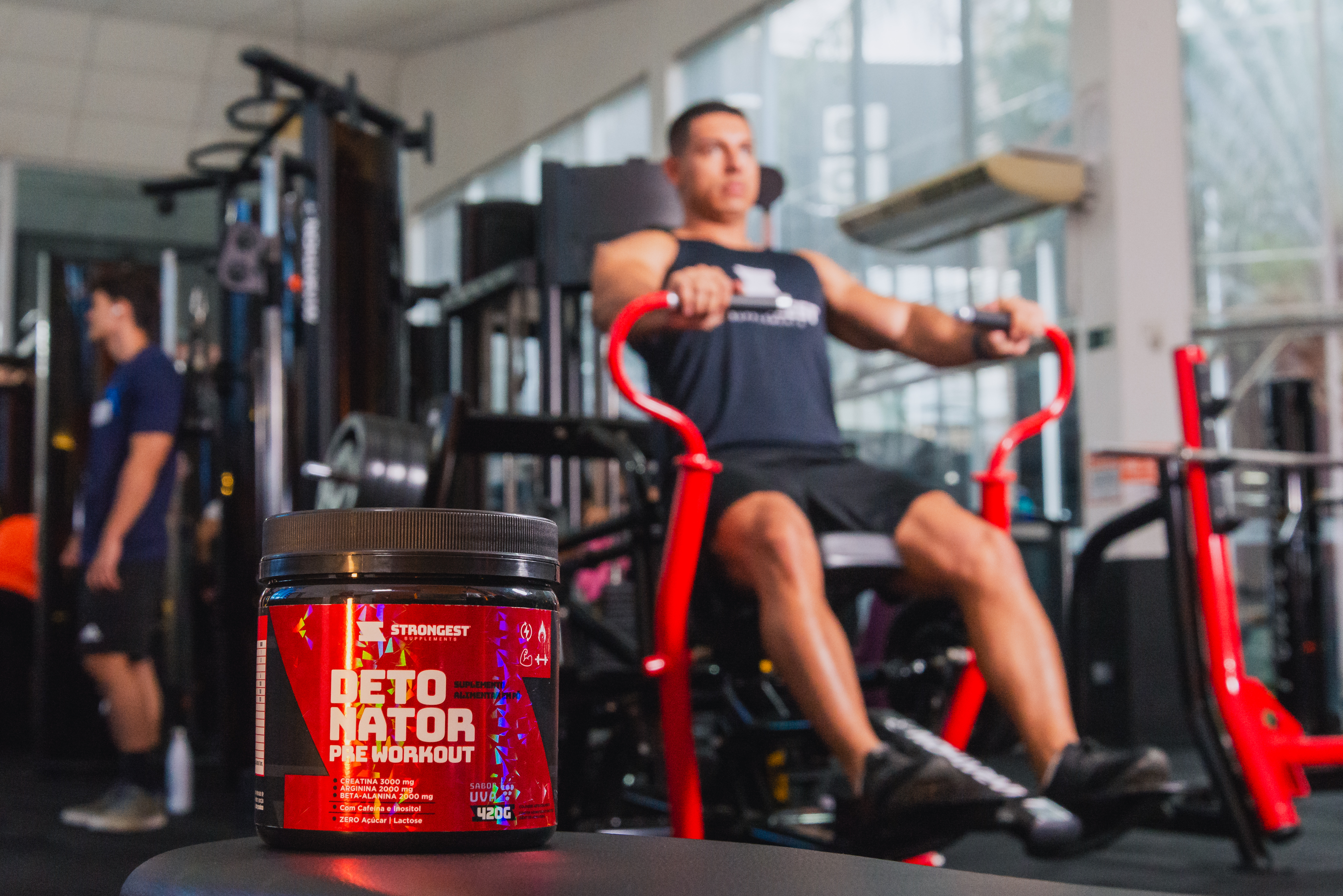 Pote do pré-treino Detonator da Strongest em primeiro plano, com homem se exercitando em uma máquina de academia ao fundo